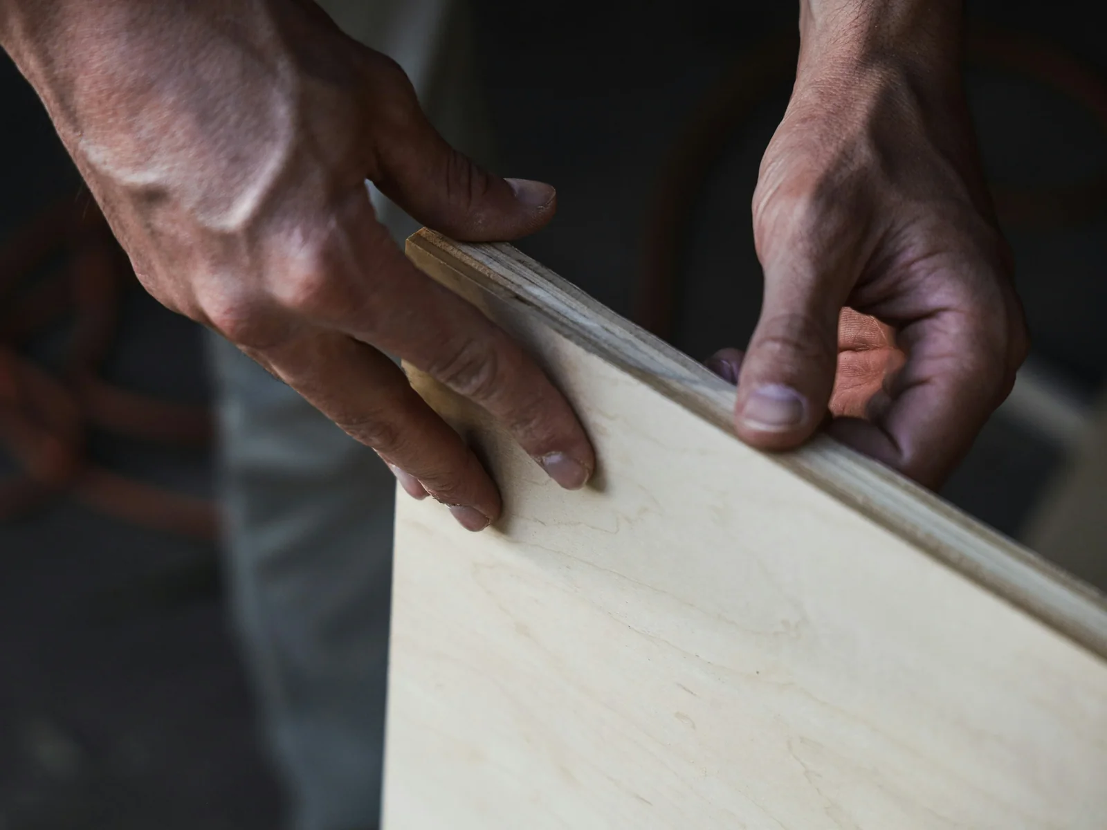 Mains d'artisan tenant le chant d'un panneau de bois clair, dans la pénombre de l'atelier