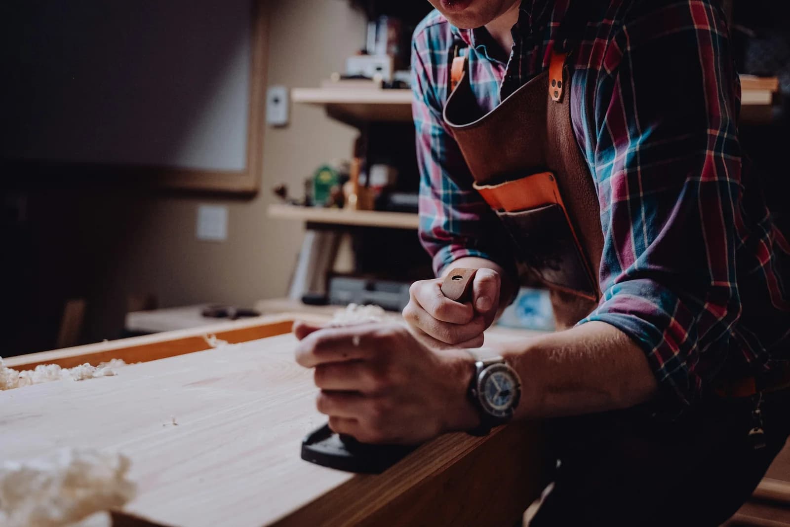 Artisan travaillant une pièce de bois à l'établi, outils rangés au mur de l'atelier